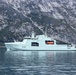 A light grey military vessel bearing hull number 435 and a Canadian maple leaf emblem is underway in calm waters against a dramatic rocky cliff backdrop. The ship appears to be a Canadian Arctic/Offshore Patrol Ship (AOPS), consistent with the Harry DeWolf-class design operated by the Royal Canadian Navy. Personnel are visible lining the upper decks, suggesting a formal or ceremonial transit.