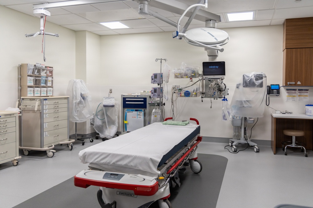 This image depicts a modern, well-equipped emergency room or trauma bay in a hospital setting. The room appears newly set up or recently cleaned, with equipment covered in protective plastic sheeting. A central emergency gurney with red accents is positioned under a large overhead surgical/examination light.