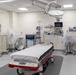 This image depicts a modern, well-equipped emergency room or trauma bay in a hospital setting. The room appears newly set up or recently cleaned, with equipment covered in protective plastic sheeting. A central emergency gurney with red accents is positioned under a large overhead surgical/examination light.