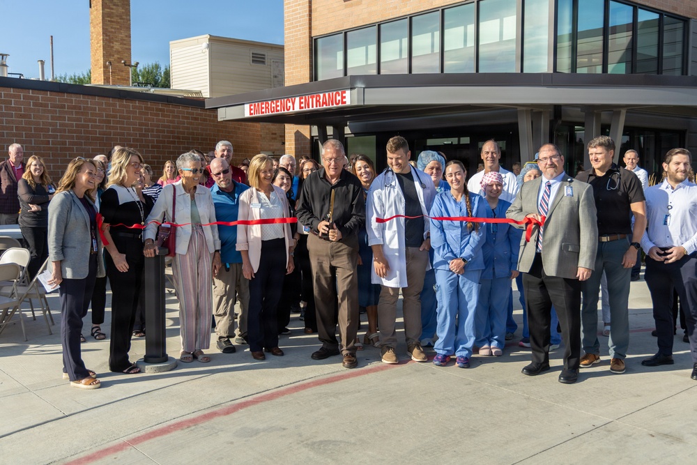 A ribbon-cutting ceremony is taking place at a hospital's newly opened or renovated Emergency Entrance. A large group of people, including medical staff in scrubs and white coats, administrators in business attire, and community members, are gathered for the event. A man in the center is cutting a red ribbon with scissors in front of the Emergency Entrance sign.