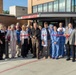 A ribbon-cutting ceremony is taking place at a hospital's newly opened or renovated Emergency Entrance. A large group of people, including medical staff in scrubs and white coats, administrators in business attire, and community members, are gathered for the event. A man in the center is cutting a red ribbon with scissors in front of the Emergency Entrance sign.