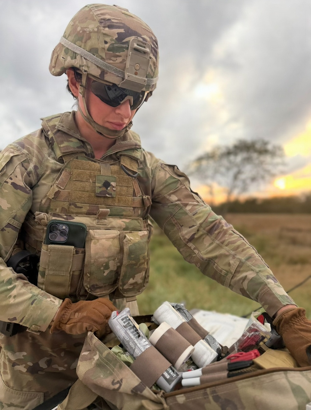 A soldier in full MultiCam/OCP camouflage uniform and tactical gear is examining or organizing medical supplies, likely from an Individual First Aid Kit (IFAK) or trauma kit. The soldier is equipped with a plate carrier, ballistic helmet with NVG mount, and tactical gloves, set against an outdoor field environment at sunset. The image appears to document a field medical readiness or training activity.