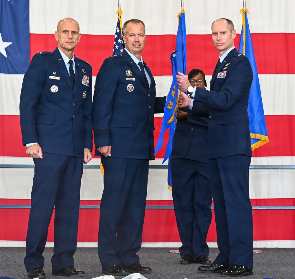 A formal U.S. Air Force change of command ceremony is taking place in front of a large American flag backdrop. Three uniformed Air Force officers are prominently featured, with two individuals exchanging a unit guidon (flag), which is the traditional centerpiece of a change of command ceremony. A fourth individual in uniform is partially visible in the background assisting with the guidon transfer.