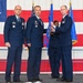 A formal U.S. Air Force change of command ceremony is taking place in front of a large American flag backdrop. Three uniformed Air Force officers are prominently featured, with two individuals exchanging a unit guidon (flag), which is the traditional centerpiece of a change of command ceremony. A fourth individual in uniform is partially visible in the background assisting with the guidon transfer.