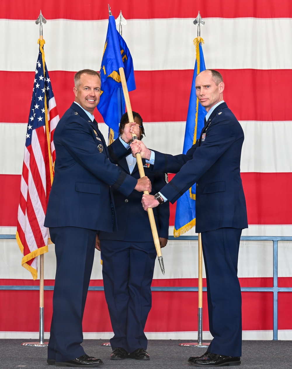 Two U.S. Air Force officers in dress blue uniforms are participating in a formal guidon/pennant passing ceremony, likely a change of command. A third uniformed airman stands between them holding the flagstaff. The ceremony takes place in front of a large American flag backdrop with additional military flags flanking the stage.