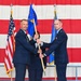 Two U.S. Air Force officers in dress blue uniforms are participating in a formal guidon/pennant passing ceremony, likely a change of command. A third uniformed airman stands between them holding the flagstaff. The ceremony takes place in front of a large American flag backdrop with additional military flags flanking the stage.