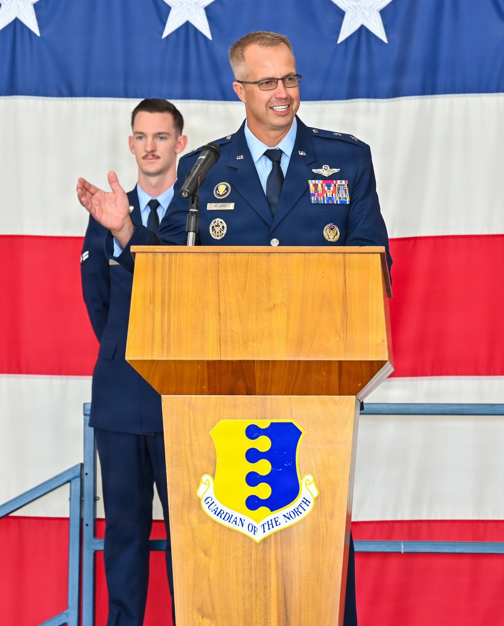 A U.S. Air Force officer in dress blues stands at a wooden podium bearing the 'Guardian of the North' shield insignia, appearing to deliver a speech or remarks at an official military ceremony. A second uniformed airman stands in the background, appearing to applaud. A large American flag serves as the backdrop.