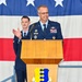 A U.S. Air Force officer in dress blues stands at a wooden podium bearing the 'Guardian of the North' shield insignia, appearing to deliver a speech or remarks at an official military ceremony. A second uniformed airman stands in the background, appearing to applaud. A large American flag serves as the backdrop.