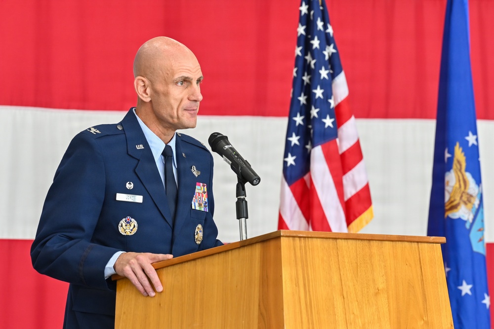 A U.S. Air Force officer in dress blues is speaking at a wooden podium with a microphone. The setting appears to be an official military ceremony, with an American flag and what appears to be an Air Force flag visible in the background. The officer displays multiple service ribbons, badges, and insignia on his uniform.