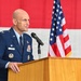 A U.S. Air Force officer in dress blues is speaking at a wooden podium with a microphone. The setting appears to be an official military ceremony, with an American flag and what appears to be an Air Force flag visible in the background. The officer displays multiple service ribbons, badges, and insignia on his uniform.