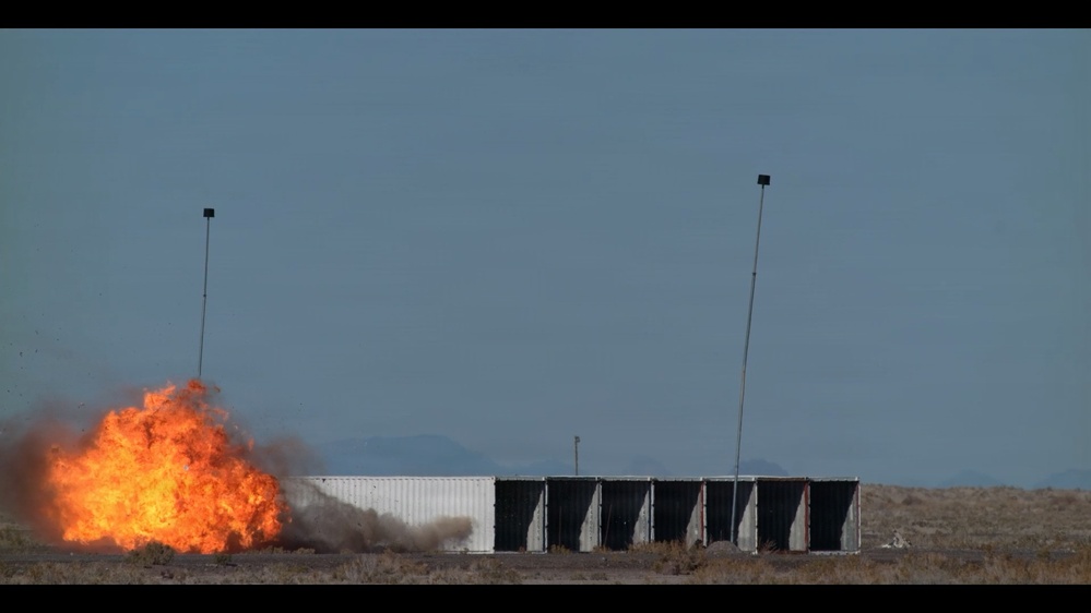 A large fireball explosion is visible on the left side of a row of shipping containers or modular structures in an open desert or arid landscape. Two tall poles with equipment mounted at their tops flank the structure, suggesting this may be a controlled test or demonstration event. The setting appears to be a remote testing range with mountains visible in the background.