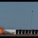 A large fireball explosion is visible on the left side of a row of shipping containers or modular structures in an open desert or arid landscape. Two tall poles with equipment mounted at their tops flank the structure, suggesting this may be a controlled test or demonstration event. The setting appears to be a remote testing range with mountains visible in the background.
