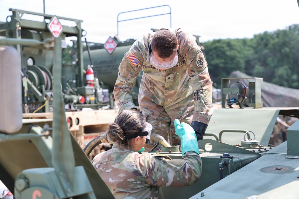 Two U.S. Army soldiers in OCP (Operational Camouflage Pattern) uniforms are performing maintenance or fueling operations on military equipment. The standing soldier, identified by a name tape reading 'ZIMMERMAN,' is leaning over to assist a second soldier who is working on what appears to be a military vehicle or fuel system component. Both soldiers are wearing protective eyewear and gloves, indicating a safety-conscious work environment.