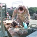 Two U.S. Army soldiers in OCP (Operational Camouflage Pattern) uniforms are performing maintenance or fueling operations on military equipment. The standing soldier, identified by a name tape reading 'ZIMMERMAN,' is leaning over to assist a second soldier who is working on what appears to be a military vehicle or fuel system component. Both soldiers are wearing protective eyewear and gloves, indicating a safety-conscious work environment.