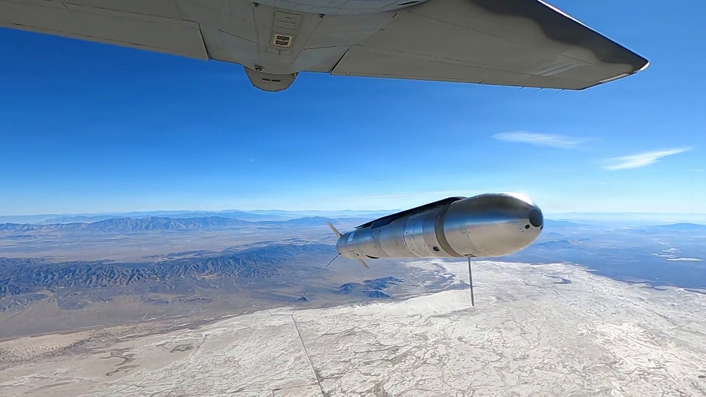 An air-launched missile or munition is captured in the moment of release from the underside of a military aircraft, photographed from a chase camera or onboard camera on the carrying aircraft. The weapon is falling away over an arid desert landscape, likely a test range environment based on the terrain features. The clear blue sky and high altitude suggest this is a controlled test or demonstration event.