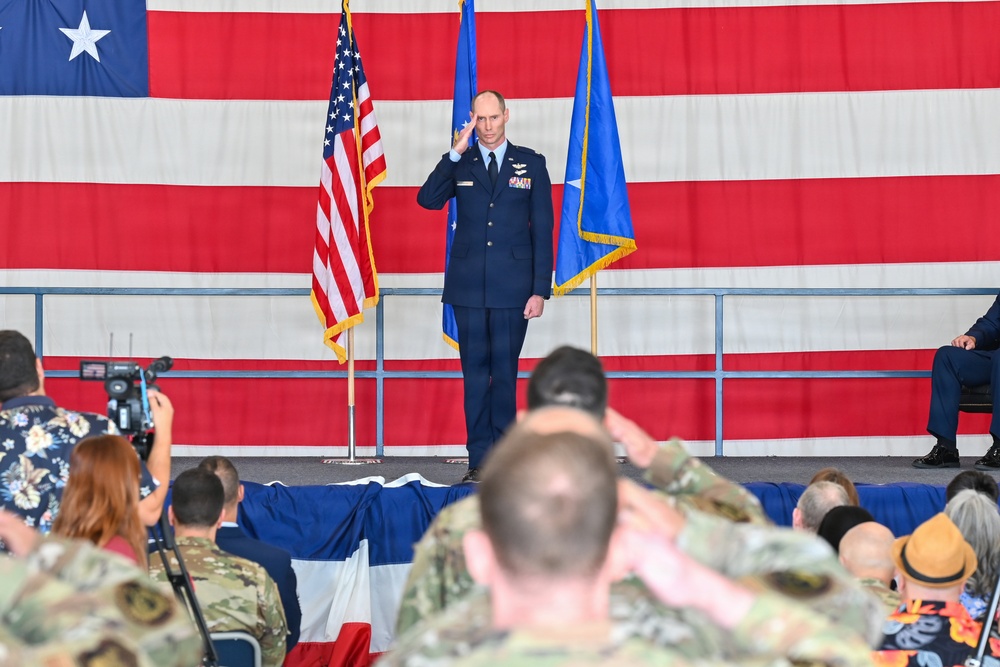 A U.S. Air Force officer in dress blues stands on a stage saluting during what appears to be a formal military ceremony, likely a change of command or activation ceremony. The backdrop features a large American flag hanging on a hangar wall, flanked by a U.S. flag and an Air Force unit flag with gold fringe. An audience of military personnel in camouflage uniforms and civilians are seated facing the stage, with some returning the salute.