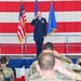A U.S. Air Force officer in dress blues stands on a stage saluting during what appears to be a formal military ceremony, likely a change of command or activation ceremony. The backdrop features a large American flag hanging on a hangar wall, flanked by a U.S. flag and an Air Force unit flag with gold fringe. An audience of military personnel in camouflage uniforms and civilians are seated facing the stage, with some returning the salute.