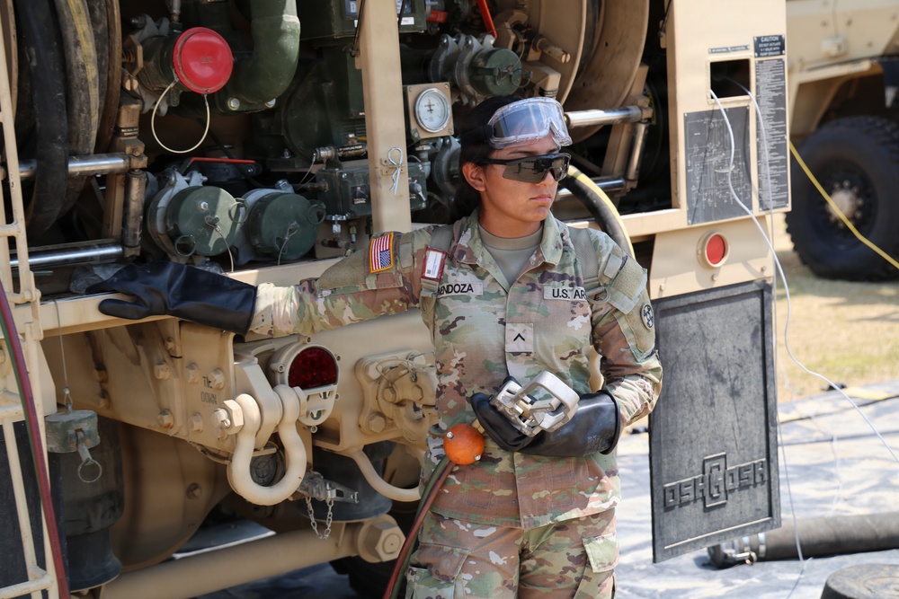 A U.S. Army soldier, identified by name tape as 'Mendoza' with the rank of Private First Class, is performing fueling or fluid transfer operations on a military tactical vehicle. The soldier is wearing full personal protective equipment including safety goggles, sunglasses, and black chemical-resistant gloves while handling a fuel nozzle or coupling connected to a hose. The vehicle visible is an Oshkosh-manufactured military truck, consistent with a fuel/water tanker or logistics support vehicle.