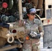 A U.S. Army soldier, identified by name tape as 'Mendoza' with the rank of Private First Class, is performing fueling or fluid transfer operations on a military tactical vehicle. The soldier is wearing full personal protective equipment including safety goggles, sunglasses, and black chemical-resistant gloves while handling a fuel nozzle or coupling connected to a hose. The vehicle visible is an Oshkosh-manufactured military truck, consistent with a fuel/water tanker or logistics support vehicle.