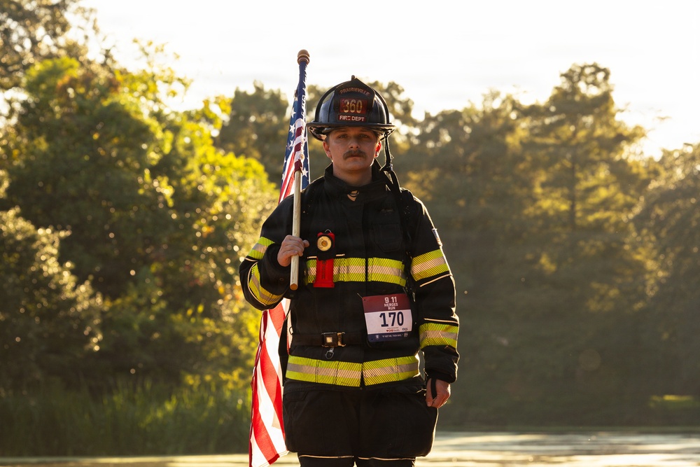 A firefighter in full turnout gear carries an American flag while participating in what appears to be a 9/11 Heroes Run event. The individual wears a black helmet marked '360' and 'Fire Dept' and displays race bib number 170. The image is taken outdoors during early morning or late afternoon light with trees visible in the background.