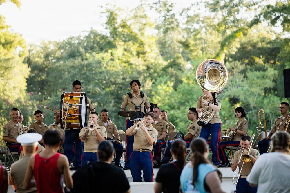 A U.S. Marine Corps band performs an outdoor concert in what appears to be a park or garden setting. Musicians in tan and blue Marine dress uniforms play a variety of brass, woodwind, and percussion instruments on a stage. A civilian audience watches from the foreground, including a person wearing a white military-style cap.