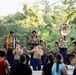 A U.S. Marine Corps band performs an outdoor concert in what appears to be a park or garden setting. Musicians in tan and blue Marine dress uniforms play a variety of brass, woodwind, and percussion instruments on a stage. A civilian audience watches from the foreground, including a person wearing a white military-style cap.