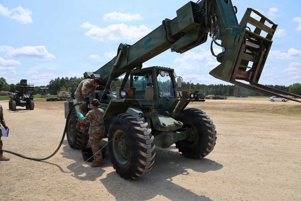 Two military personnel in camouflage uniforms are performing maintenance or fueling operations on a military rough-terrain forklift (likely an RT-series or similar telehandler) painted in olive drab green. The vehicle is positioned on a sandy, open training area with additional military vehicles and tree line visible in the background. One soldier is positioned on top of the vehicle while another works at ground level with a hose.