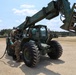Two military personnel in camouflage uniforms are performing maintenance or fueling operations on a military rough-terrain forklift (likely an RT-series or similar telehandler) painted in olive drab green. The vehicle is positioned on a sandy, open training area with additional military vehicles and tree line visible in the background. One soldier is positioned on top of the vehicle while another works at ground level with a hose.