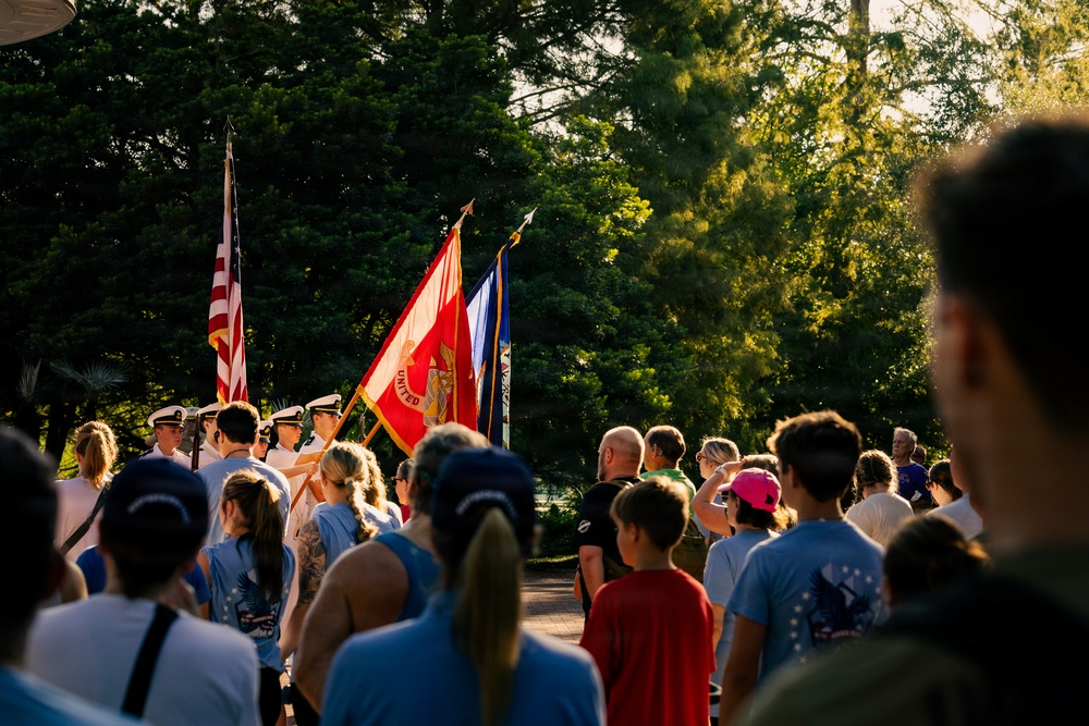 A military color guard ceremony is taking place outdoors, likely at a patriotic or memorial event. Uniformed personnel are carrying multiple flags including the American flag and what appears to be a U.S. Marine Corps flag, while a crowd of civilians watches. The scene is bathed in warm golden-hour sunlight with lush green trees in the background.