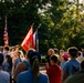 A military color guard ceremony is taking place outdoors, likely at a patriotic or memorial event. Uniformed personnel are carrying multiple flags including the American flag and what appears to be a U.S. Marine Corps flag, while a crowd of civilians watches. The scene is bathed in warm golden-hour sunlight with lush green trees in the background.