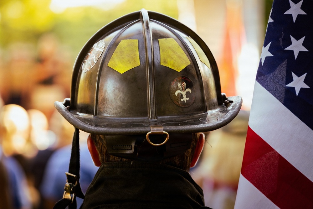 A close-up rear view of a firefighter wearing a traditional dark-colored fire helmet with yellow reflective markings and a fleur-de-lis emblem sticker. An American flag is visible to the right side of the frame. The background is blurred, suggesting a public gathering or ceremony.