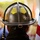 Audubon Park Hosts the 2026 New Orleans 9/11 Heroes Run A close-up rear view of a firefighter wearing a traditional dark-colored fire helmet with yellow reflective markings and a fleur-de-lis emblem sticker. An American flag is visible to the right side of the frame. The background is blurred, suggesting a public gathering or ceremony.