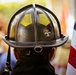A close-up rear view of a firefighter wearing a traditional dark-colored fire helmet with yellow reflective markings and a fleur-de-lis emblem sticker. An American flag is visible to the right side of the frame. The background is blurred, suggesting a public gathering or ceremony.