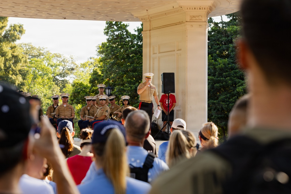 A U.S. Marine Corps officer addresses a crowd from a white neoclassical bandstand or pavilion, with a military band seated behind him in tan service uniforms. The outdoor ceremony or performance appears to be taking place in a park-like setting with lush green trees in the background. A civilian audience watches from the foreground, with some individuals photographing the event.