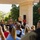 Audubon Park Hosts the 2026 New Orleans 9/11 Heroes Run A U.S. Marine Corps officer addresses a crowd from a white neoclassical bandstand or pavilion, with a military band seated behind him in tan service uniforms. The outdoor ceremony or performance appears to be taking place in a park-like setting with lush green trees in the background. A civilian audience watches from the foreground, with some individuals photographing the event.