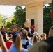 A U.S. Marine Corps officer addresses a crowd from a white neoclassical bandstand or pavilion, with a military band seated behind him in tan service uniforms. The outdoor ceremony or performance appears to be taking place in a park-like setting with lush green trees in the background. A civilian audience watches from the foreground, with some individuals photographing the event.