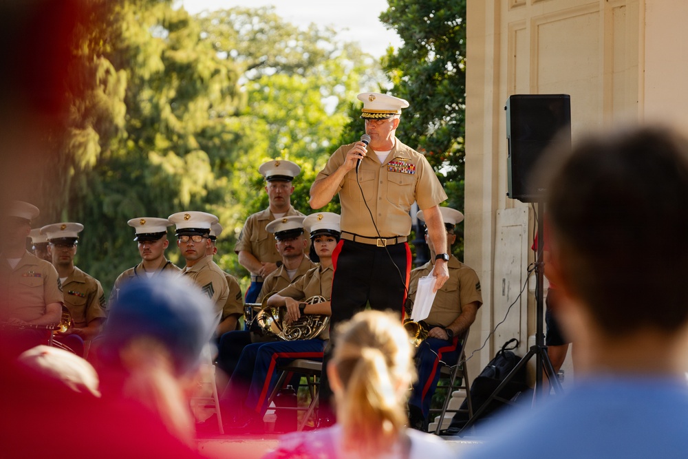 A senior U.S. Marine Corps officer is addressing an audience via microphone at what appears to be an outdoor military band concert or ceremony. He is standing before a seated ensemble of uniformed Marines, several of whom are holding brass instruments. The setting appears to be a formal outdoor venue with a classical architectural backdrop and lush trees.