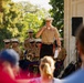 A senior U.S. Marine Corps officer is addressing an audience via microphone at what appears to be an outdoor military band concert or ceremony. He is standing before a seated ensemble of uniformed Marines, several of whom are holding brass instruments. The setting appears to be a formal outdoor venue with a classical architectural backdrop and lush trees.