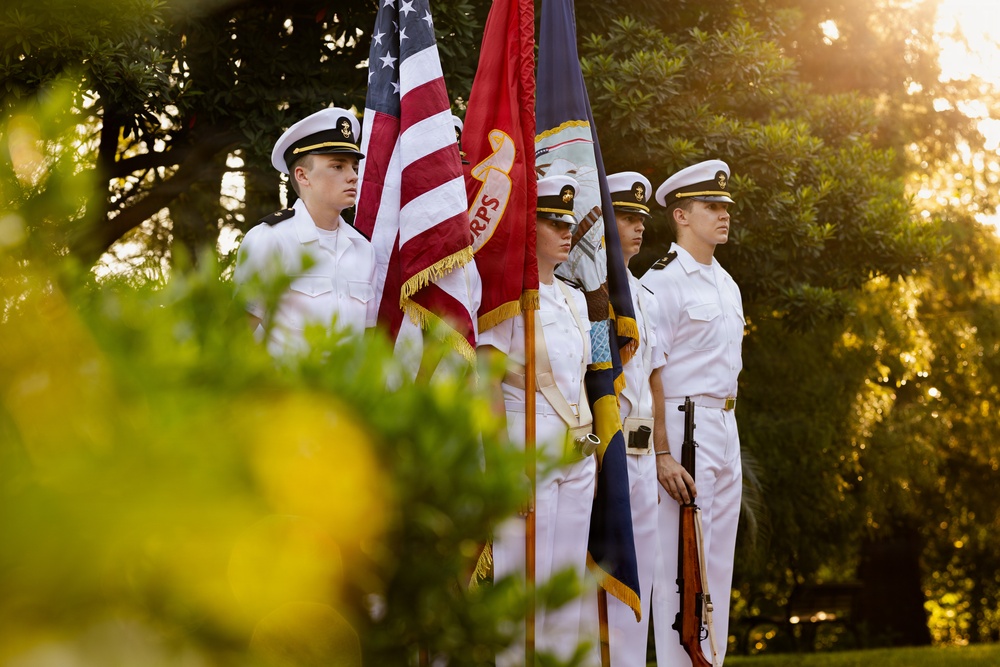 A military color guard of four personnel dressed in white naval uniforms stands at attention during a formal ceremony. They are holding multiple flags including the United States flag, a red flag with 'RPS' visible, and a dark blue flag. The scene is photographed outdoors during golden hour, with lush green foliage framing the composition.