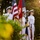 Audubon Park Hosts the 2026 New Orleans 9/11 Heroes Run A military color guard of four personnel dressed in white naval uniforms stands at attention during a formal ceremony. They are holding multiple flags including the United States flag, a red flag with 'RPS' visible, and a dark blue flag. The scene is photographed outdoors during golden hour, with lush green foliage framing the composition.