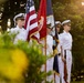 A military color guard of four personnel dressed in white naval uniforms stands at attention during a formal ceremony. They are holding multiple flags including the United States flag, a red flag with 'RPS' visible, and a dark blue flag. The scene is photographed outdoors during golden hour, with lush green foliage framing the composition.
