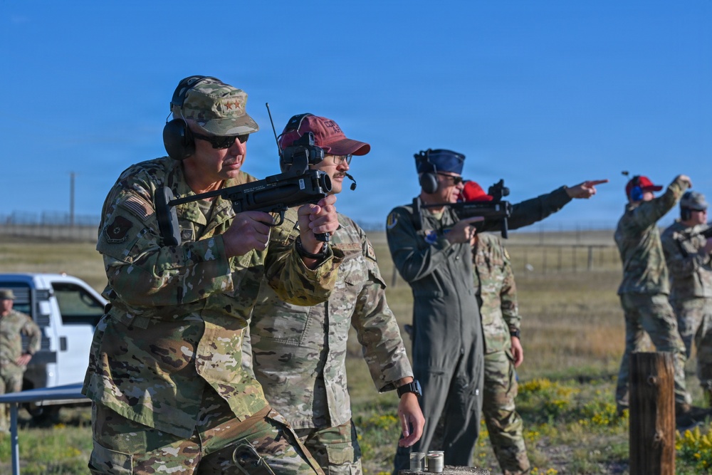 Military personnel are participating in an outdoor shooting range exercise or training event. Multiple individuals in camouflage uniforms and hearing protection are handling firearms, with at least one person actively aiming a weapon. The setting appears to be an open, flat terrain consistent with a military training range.