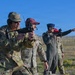 Military personnel are participating in an outdoor shooting range exercise or training event. Multiple individuals in camouflage uniforms and hearing protection are handling firearms, with at least one person actively aiming a weapon. The setting appears to be an open, flat terrain consistent with a military training range.