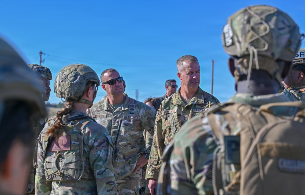 A group of U.S. Air Force personnel in OCP (Operational Camouflage Pattern) uniforms are gathered outdoors in what appears to be a briefing or after-action review. Several individuals are wearing tactical helmets and body armor, while at least two senior personnel without helmets appear to be addressing the group. The setting appears to be an open field or training area under clear blue skies.