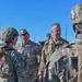 A group of U.S. Air Force personnel in OCP (Operational Camouflage Pattern) uniforms are gathered outdoors in what appears to be a briefing or after-action review. Several individuals are wearing tactical helmets and body armor, while at least two senior personnel without helmets appear to be addressing the group. The setting appears to be an open field or training area under clear blue skies.