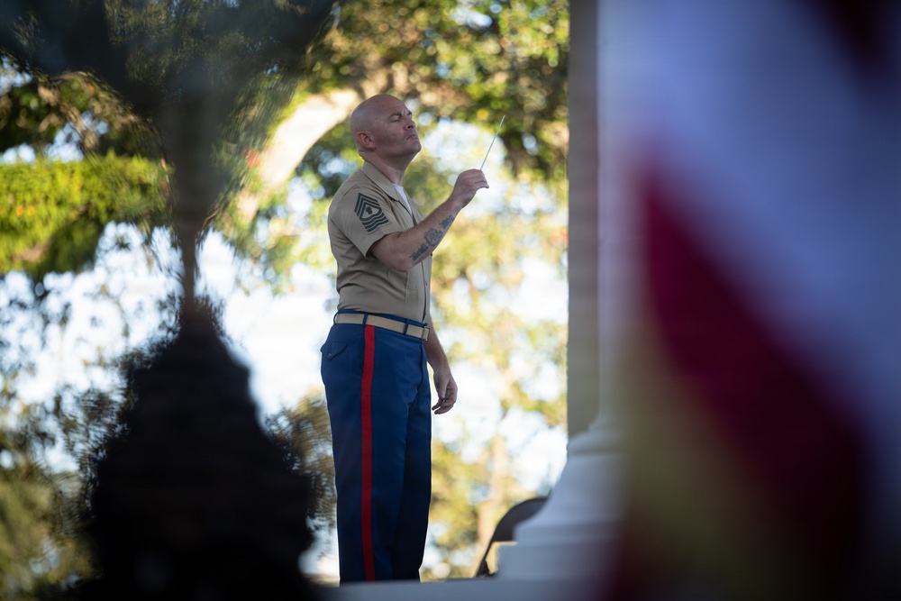 A U.S. Marine in dress uniform is conducting a musical performance outdoors, holding a conductor's baton raised in his right hand. He appears to be a senior non-commissioned officer based on the rank insignia visible on his collar. An American flag is partially visible in the foreground right, and the scene is framed by blurred foliage and architectural elements.