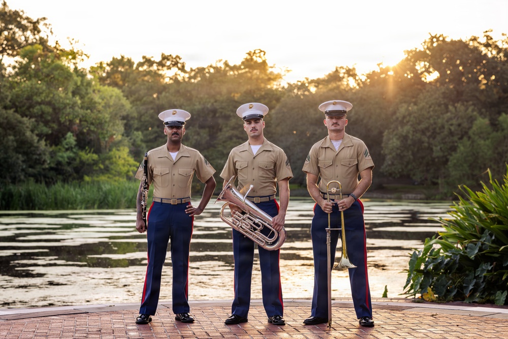 Three U.S. Marine Corps musicians in service uniforms (tan shirts, blue trousers with red stripe) pose for a formal portrait at golden hour near a reflective pond. Each Marine holds a different instrument, suggesting they are members of a military band unit. The setting is an outdoor garden or park environment with lush greenery and warm sunset lighting.