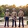 Audubon Park Hosts the 2026 New Orleans 9/11 Heroes Run Three U.S. Marine Corps musicians in service uniforms (tan shirts, blue trousers with red stripe) pose for a formal portrait at golden hour near a reflective pond. Each Marine holds a different instrument, suggesting they are members of a military band unit. The setting is an outdoor garden or park environment with lush greenery and warm sunset lighting.