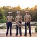 Three U.S. Marine Corps musicians in service uniforms (tan shirts, blue trousers with red stripe) pose for a formal portrait at golden hour near a reflective pond. Each Marine holds a different instrument, suggesting they are members of a military band unit. The setting is an outdoor garden or park environment with lush greenery and warm sunset lighting.