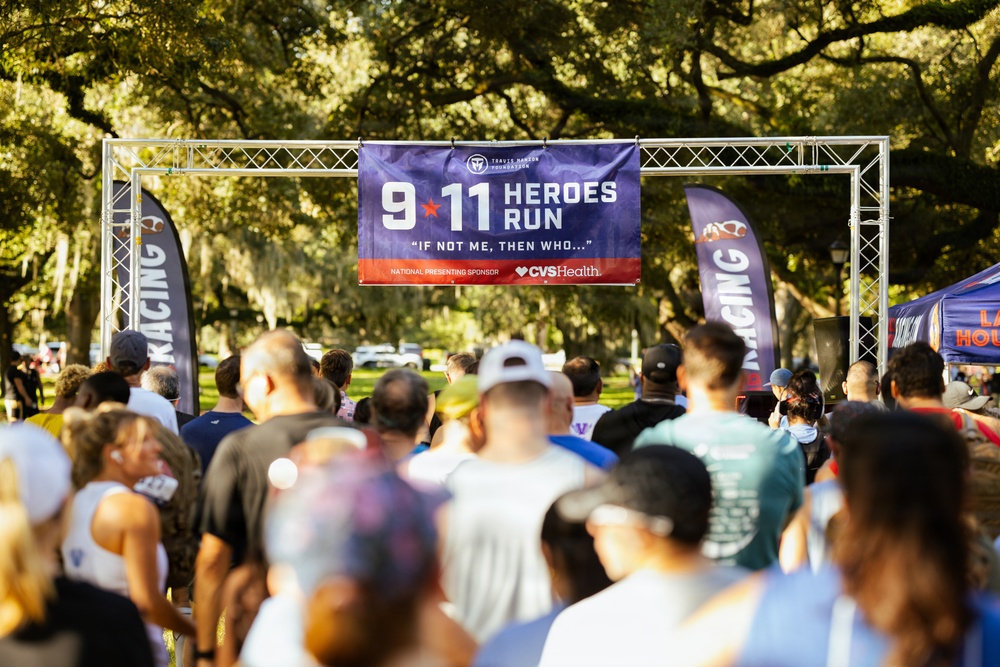 A large crowd of participants is gathered at the starting area of the '9/11 Heroes Run,' an annual commemorative race organized by the Travis Manion Foundation. The event is held outdoors in a park-like setting with large oak trees providing shade. CVS Health is identified as the national presenting sponsor on the main banner.